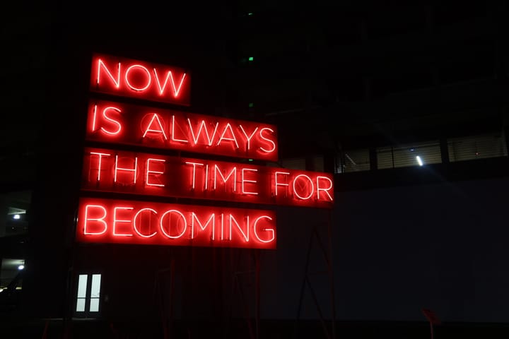 A red neon sign with the quote "Now is always the time for becoming"